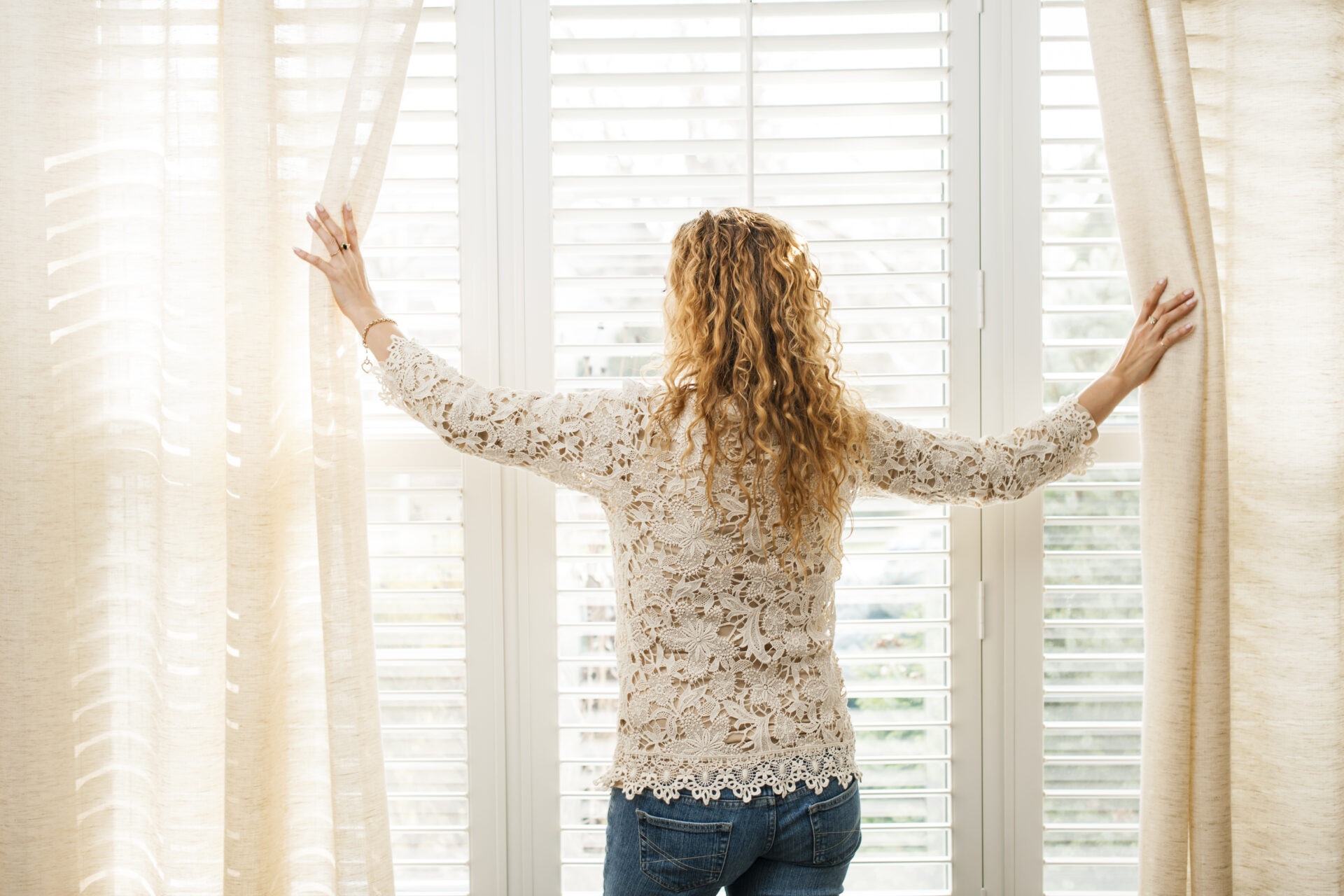 Woman looking out California blinds. Custom blinds for any size window at Wholesale Blind Factory.